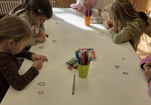 Dzieci siedzą przy stole i kolorują małe papierowe elementy podczas warsztatów plastycznych