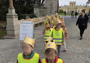 Przedszkolaki w odblaskowych kamizelkach i papierowych koronach spacerują alejką przed Pałacem w Wilanowie.