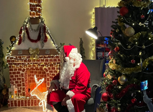 Święty Mikołaj siedzący w fotelu przy kominku i choince.