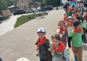 Grupa dzieci ubranych w pomarańczowe chustki, które idą w pochodzie ulicznym, uśmiechnięte i trzymające się za ręce, niektóre z nich mają dekoracje z liści i kwiatów.