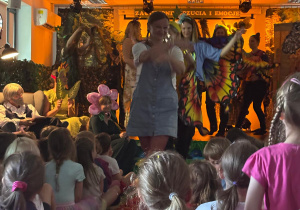Występ teatralny dla dzieci, na scenie osoby dorosłe przebrane są za kolorowe motyle, kwiaty i rośliny, a w tle widnieje napis „Zagubione uczucia i emocje”. Przed sceną siedzi liczna grupa dzieci, z zaciekawieniem oglądając przedstawienie.