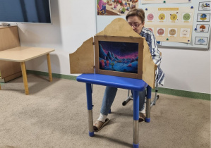 Pani Beatka siedzącą w sali przedszkolnej i prezentującą dzieciom teatrzyk kamishibai, w którym pokazuje kolorowe ilustracje z bajki.