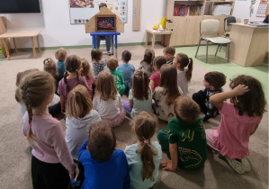 Grupa dzieci siedzących na dywanie w sali przedszkolnej i wspólnie oglądających teatrzyk kamishibai, w którym prezentowane są kolorowe ilustracje bajki.