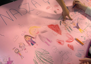 duży różowy plakat z napisem „Nasza Drużyna”, na którym dzieci rysują kredkami postacie, serca i różne symbole, wspólnie tworząc pracę plastyczną.