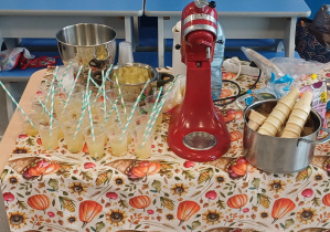 Na zdjęciu widać stół przykryty jesiennym obrusem z motywami dyń, liści i słoneczników. Na stole stoją plastikowe kubeczki z żółtym napojem i słomkami, metalowe miski z pokrojonymi owocami, czerwony mikser planetarny oraz garnek z pokrojonymi bananami. Przygotowano stanowisko do robienia koktajli owocowych lub smoothie podczas zajęć kulinarnych dla dzieci.