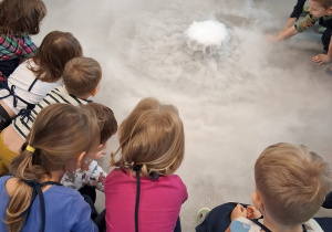 Na zdjęciu widać grupę dzieci siedzących w kręgu wokół naczynia, z którego unosi się gęsta mgła przypominająca dym. Dzieci w fartuszkach z zainteresowaniem obserwują i dotykają pary.