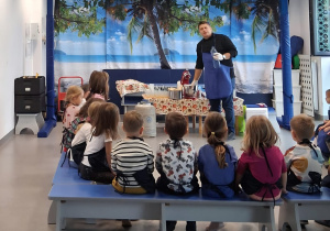 Na zdjęciu widać grupę dzieci siedzących na ławkach, zwróconych w stronę prowadzącego zajęcia mężczyzny w niebieskim fartuchu. Na stole przed nim znajdują się różne przyrządy i składniki do warsztatów. W tle widoczna jest dekoracja przedstawiająca plażę z palmami i morzem. Dzieci uważnie obserwują pokaz edukacyjny.