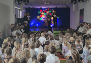 Widok z tyłu sali podczas uroczystości przedszkolnej. Na scenie stoją nauczyciele, a przed nimi siedzi duża grupa dzieci w odświętnych strojach. Tło sceny zdobi kolorowe drzewo z napisem „Święto Edukacji Narodowej”.