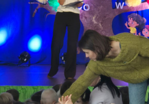 Na scenie stoi prowadząca uroczystość z okazji Dnia Edukacji Narodowej. Za nią dekoracja przedstawiająca drzewo z kolorowych bibułowych kwiatów. Przed sceną siedzą dzieci.