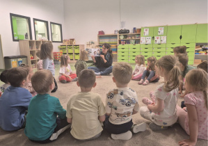 Na zdjęciu widać kobietę siedzącą na dywanie w przedszkolu, która czyta książkę grupie dzieci siedzących w kręgu. Dzieci uważnie słuchają i obserwują ilustracje w książce. W tle widać zielone szafki i zabawki w kąciku zabaw.