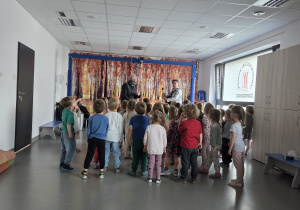 Grupa przedszkolaków stojących w sali i obserwujących dwóch mężczyzn prowadzących pokaz muzyczny na tle jesiennej dekoracji przedstawiającej las.