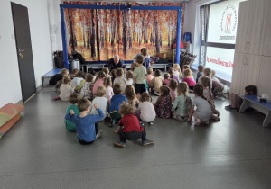 Grupa dzieci siedzących na podłodze w sali, które oglądają występ prowadzone przez dwóch mężczyzn na tle jesiennej dekoracji przedstawiającej las.