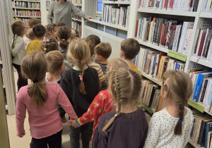 Grupa dzieci stojących w rzędzie między regałami pełnymi książek w bibliotece, słuchających kobiety, która opowiada im o zbiorach bibliotecznych i zasadach korzystania z biblioteki.