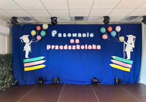 Uroczystość w przedszkolu,dzieci siedzą na kolorowym dywanie nauczycielka przemawia na scenie ozdobionej napisem „Pasowanie na przedszkolaka”.