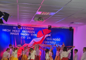 dzieci występujące na scenie podczas uroczystości z okazji 11 listopada, ubrane w białe koszule i kolorowe spódnice lub spodnie, tańczą z biało-czerwonymi wstążkami na tle dekoracji patriotycznej z napisem o bohaterach, którzy walczyli o wolność.