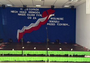 Scena udekorowana z okazji Święta Niepodległości - tło stanowi granatowa kurtyna z napisem „11 listopada Niech hołd oddadzą nasze głosy tym, co za wolność odeszli przed czasem…”, a centralnym elementem jest biało-czerwona wstęga ułożona w kształt orła.