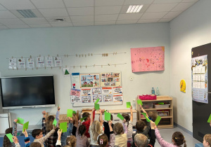 Grupa przedszkolaków siedzących na dywanie i trzymających w górze zielone kartoniki podczas zajęć prowadzonych przy tablicy edukacyjnej w sali przedszkolnej.