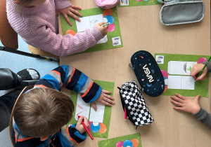 Dzieci siedzące przy stoliku i rysujące na kartach pracy zapamiętane prawa dziecka ,obok których leżą piórniki.