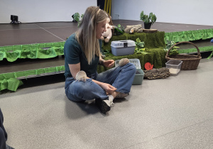 Prowadząca warsztaty kobieta siedzącą na podłodze w sali, która podczas zajęć pokazuje dzieciom jeże, trzyma jednego na kolanach, a obok niej stoją pojemniki i dekoracje przypominające leśne otoczenie.