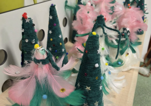 Świąteczne choinki wykonane przez dzieci podczas przedszkolnych warsztatów plastycznych, ozdobione piórkami, pomponami i gwiazdkami.