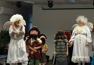 Sceną, na której stoją dorośli przebrani za anioły oraz dzieci w kolorowych, bajkowych kostiumach, a przed sceną na podłodze siedzi duża grupa przedszkolaków uważnie oglądających przedstawienie.