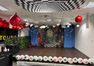 Na zdjęciu widać scenę w przedszkolu udekorowaną w stylu „Alicji w Krainie Czarów” – z czarno-białą szachownicą na suficie, czerwonymi balonami w kształcie serc, białymi chmurkami oraz tłem z bajkowym ogrodem i motywami kart do gry.