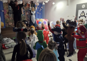 Na zdjęciu widać bal karnawałowy w sali przedszkolnej – dzieci w kolorowych przebraniach bawią się i tańczą, unosząc ręce do góry, a na scenie animatorzy prowadzą zabawę z balonami pod dekoracjami w biało-czarną szachownicę i motyw kart.