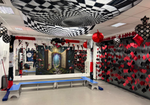 Na zdjęciu widać przedszkolną salę udekorowaną w stylu „Alicji w Krainie Czarów” – z czarno-białą szachownicą na suficie, czerwono-czarnymi sercami i symbolami kart na ścianach oraz dużym tłem z otwartą księgą i magicznym przejściem.