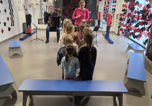 Grupa przedszkolaków stojących naprzeciwko sceny, na której kobieta prowadzi zajęcia muzyczne, a siedzący obok mężczyzna gra na akordeonie, w sali udekorowanej bajkowymi motywami kart do gry i kolorowymi dekoracjami.