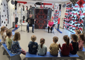 Grupa przedszkolaków siedzących na ławkach i słuchających występu muzycznego - mężczyzna gra na akordeonie, a kobieta śpiewa – w sali udekorowanej motywami kart do gry i bajkowej scenografii.