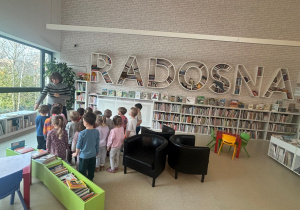 Dzieci siedzą w bibliotece wokół osoby prowadzącej zajęcia, słuchając i oglądając książki w kolorowym kąciku czytelniczym.