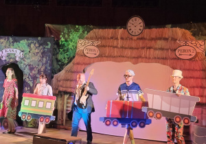 Na zdjęciu widać scenę teatralną, na której aktorzy w kolorowych kostiumach, trzymając makiety wagonów, odgrywają dynamiczną scenę na tle dekoracji przedstawiającej peron kolejowy.