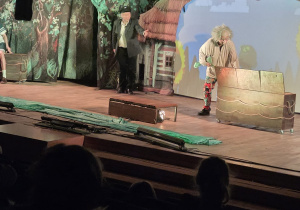 Na zdjęciu widać scenę teatralną, na której aktorzy w kostiumach odgrywają przedstawienie na tle dekoracji przypominającej stację kolejową z kasą i peronem, a widzowie obserwują spektakl z sali.