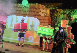 Na zdjęciu widać scenę teatralną, na której aktorzy w kolorowych kostiumach odgrywają przedstawienie związane z podróżą pociągiem, trzymając makiety wagonów na tle dekoracji przypominającej peron kolejowy.