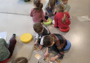 Na zdjęciu widać dzieci siedzące na podłodze w sali przedszkolnej, które wspólnie układają obrazkowe puzzle, podczas gdy nauczycielka pomaga im w zadaniu.