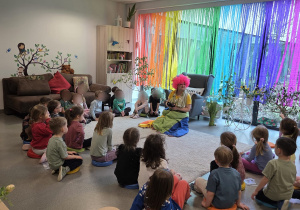 Na zdjęciu widać dzieci siedzące w kręgu w sali przedszkolnej, które z uwagą słuchają osoby prowadzącej zajęcia w kolorowym stroju na tle tęczowej dekoracji przy oknie.