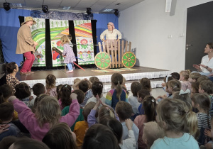 Grupa dzieci siedzących na podłodze i oglądających przedstawienie teatralne na scenie, gdzie aktorzy oraz dwoje dzieci biorą udział w barwnej, interaktywnej scenie z dekoracją przedstawiającą wiejskie tło i drewniany wóz.