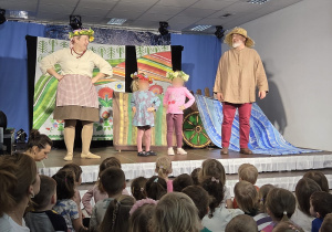 Dzieci siedzące przed sceną i oglądające przedstawienie, w którym aktorzy w strojach ludowych oraz dwoje dzieci uczestniczą w barwnej, interaktywnej scenie z dekoracją przedstawiającą wiejskie krajobrazy i wóz.