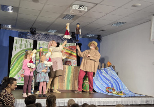 Dzieci siedzące przed sceną i oglądające przedstawienie, podczas którego aktorzy oraz kilkoro dzieci trzymają kukiełki i wspólnie odgrywają barwną scenę na tle dekoracji przedstawiającej wiejski krajobraz.