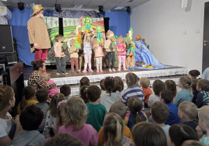 Dzieci siedzące przed sceną i oglądające przedstawienie, podczas którego grupa dzieci wraz z aktorami stoi na scenie i trzyma kolorowe kukiełki, odgrywając wspólną, radosną scenę.