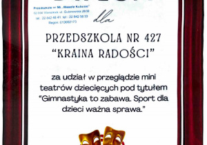 Na zdjęciu widoczny jest dyplom w dekoracyjnej, teatralnej oprawie z motywem kurtyny i masek, przyznany Przedszkolu nr 427 „Kraina Radości” za udział w przeglądzie mini teatrów dziecięcych pt. „Gimnastyka to zabawa. Sport dla dzieci ważna sprawa.”.