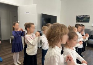Dzieci podczas wspólnego występu - stoją w półokręgu, ubrane elegancko (białe bluzki, granatowe spódnice i spodnie), wykonują gesty rękami i klaszczą, śpiewają i prezentują przygotowany układ dla gości.