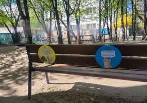 Ławka w przedszkolnym ogrodzie z zamocowanymi kolorowymi kartami zadaniowymi, które stanowią element ścieżki edukacyjnej dla dzieci.