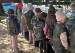 Grupa dzieci podczas zajęć na świeżym powietrzu, które w ochronnych pelerynach z worków foliowych obserwują i uczestniczą w aktywności przy stanowisku edukacyjnym