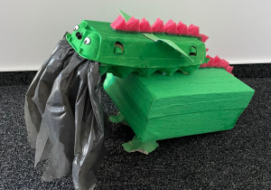 Praca plastyczna przedstawiająca zielonego eko-smoka wykonanego z materiałów recyklingowych, który „pożera” plastikową torbę.