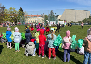 Dzieci uczestniczące w plenerowym wydarzeniu z okazji Dnia Ziemi, które oglądają występ na boisku, trzymając zielone kwiatki