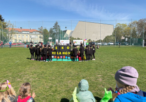 Dzieci biorące udział w plenerowym wydarzeniu ekologicznym, które prezentują wspólny występ z napisem „EKO WILANÓW!”.