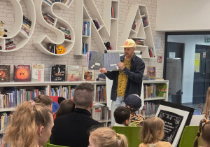 Dzieci wraz z rodzicami uczestniczą w spotkaniu autorskim w bibliotece, podczas którego prowadzący prezentuje książkę i czyta jej fragmenty zgromadzonej publiczności.