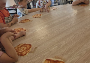 Dzieci uczestniczące w warsztatach kulinarnych, podczas których samodzielnie smarowały sosem pizzę, poznając etapy przygotowywania pizzy i rozwijając swoje umiejętności manualne.