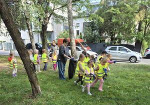 Dzieci wraz z nauczycielkami sprzątają teren zielony, zbierając znalezione śmieci do worka.